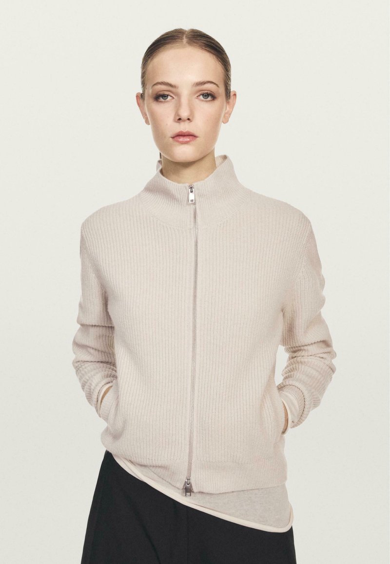 Maglione a coste beige chiaro con colletto alto, zip frontale completa e maniche lunghe, caratterizzato da una texture morbida e una silhouette aderente.