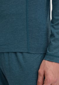 Teal overhemd met lange mouwen en ronde hals; heeft platte naden en een gladde textuur, gecombineerd met bijpassende broek. Close-up van de mouw en de zijkant.