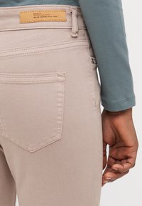 Jeans de mezclilla beige claro con bolsillos traseros cosidos, diseño ajustado y una etiqueta marrón en la cinturilla. Textura ligeramente desgastada y acabado suave.