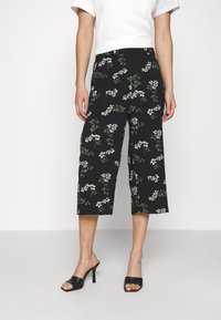 Culottes noires avec un motif floral comprenant des fleurs blanches et des feuilles vertes. Fabriquées dans un tissu léger, avec une coupe large et une longueur raccourcie.