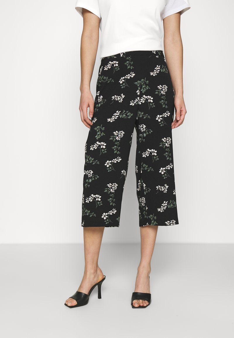 Culottes noires avec un motif floral comprenant des fleurs blanches et des feuilles vertes. Fabriquées dans un tissu léger, avec une coupe large et une longueur raccourcie.