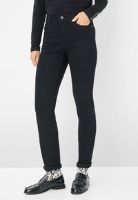 Jeans neri slim-fit con una texture liscia, dotati di tasche frontali, chiusura a bottone e orlo pulito, abbinati a calzini con motivo e scarpe nere.