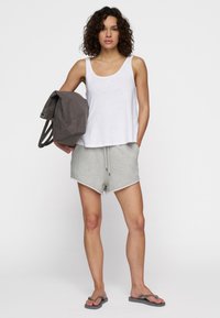 Top sin mangas blanco, pantalones cortos grises con cordón y chanclas; modelo sosteniendo una mochila gris, con un diseño minimalista y texturas suaves.