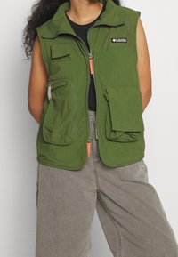 Zelená bezrukávová vesta s několika kapsami, zapínáním na zip s oranžovými akcenty a síťovinovými detaily na ramenou. Nositá přes černý tílko.