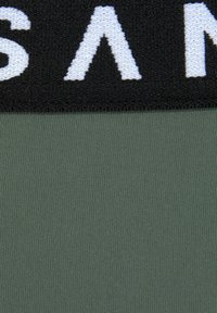 Tessuto verde oliva con cintura nera testurizzata caratterizzata da lettering bianco a contrasto. Materiale liscio e elasticizzato con un motivo a coste.