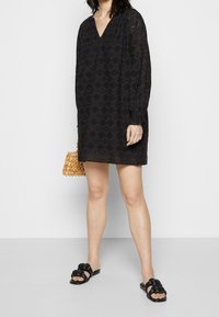 Donna con un mini abito lungo a maniche nere e fantasia, che tiene in mano una borsa a tracolla di piccole perline di legno e sandali neri flat intrecciati.