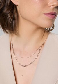 Naszyjnik warstwowy z dwoma łańcuchami; jeden z wydłużonymi srebrnymi ogniwami, a drugi z delikatnego srebrnego łańcucha, oba leżące na jasnym tkaninowym topie.