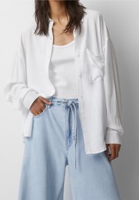 Camicia bianca oversize con bottoni, realizzata in tessuto leggero, abbinata a una canotta bianca aderente e jeans a gamba larga blu chiaro con cordoncino.