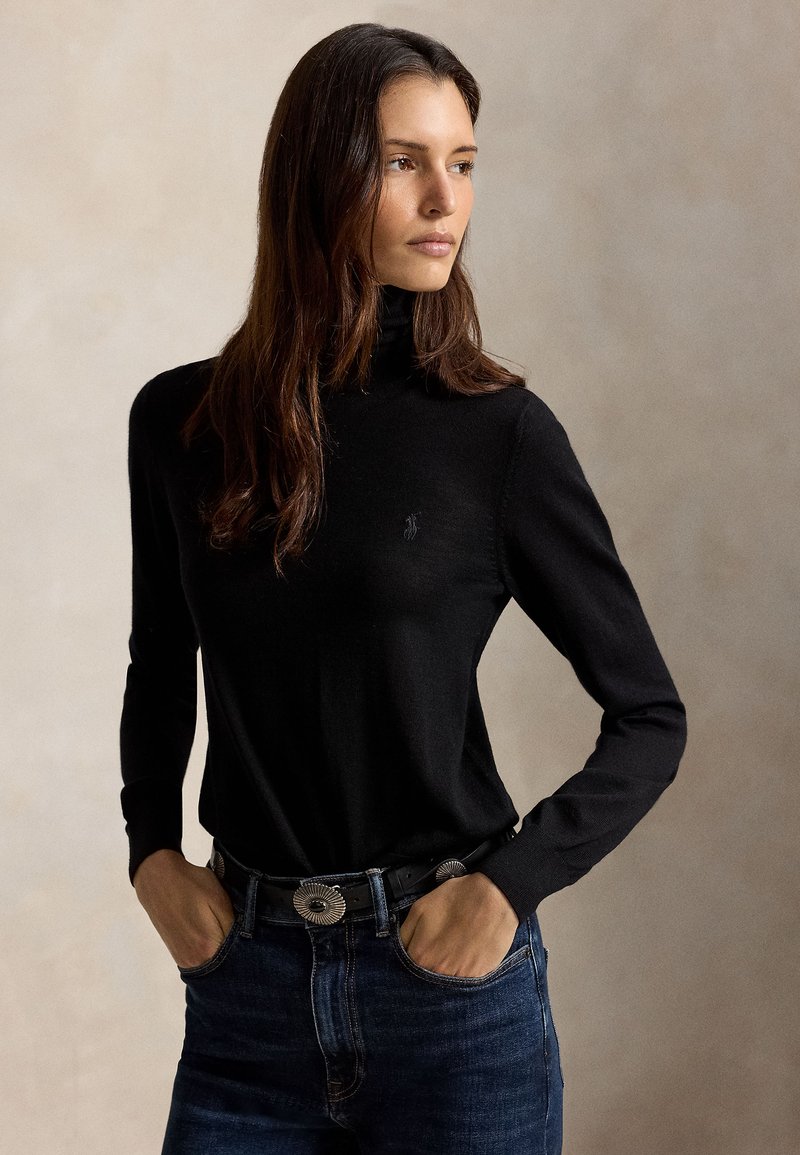 Femme aux longs cheveux bruns portant un pull à col roulé noir, un jean foncé et une ceinture avec de grands conchos argentés, mains dans les poches, regardant vers la droite.