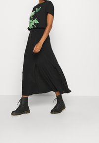 Camiseta negra con diseño gráfico verde, combinada con una falda maxi negra con aberturas laterales. Lleva puestas botas cortas negras con cordones.
