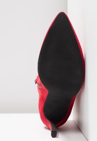 Scarpa alta in suede rosso con punta affilata e suola nera texturizzata. Dettaglio della zip laterale visibile sull'esterno della scarpa.