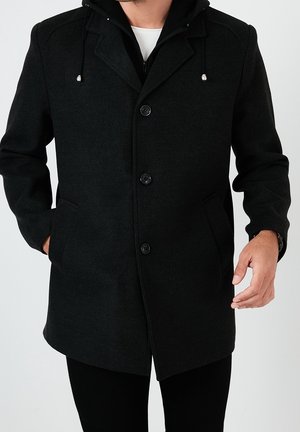 Mann trägt einen schwarzen Kapuzenmantel aus Wolle mit drei Knöpfen, Seitentaschen, die Hände teilweise in den Taschen, darüber ein weißes Hemd und schwarze Hose.