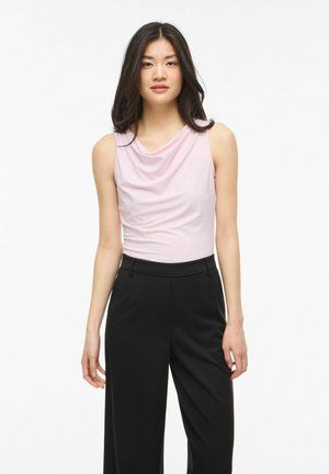 Kobieta w bezrękawnikowym jasnoróżowym marszczonym topie i czarnych szerokich spodniach z wysokim stanem stoi na białym tle.