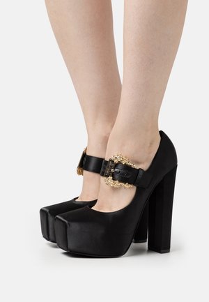 Svarta högklackade platåskor med rund tå, tillverkade av slätt läder. Har en dekorativ guldspänne på ankelselen.