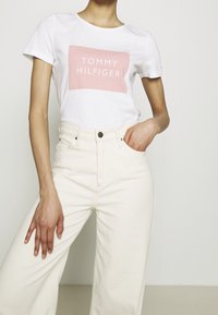 Camiseta blanca de algodón con un logo rectangular rosa que dice "TOMMY HILFIGER". Combinada con pantalones de pierna ancha color crema de cintura alta.