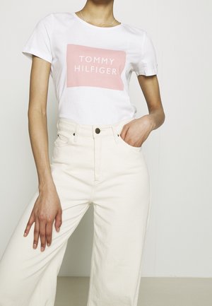 Camiseta blanca de algodón con un logo rectangular rosa que dice "TOMMY HILFIGER". Combinada con pantalones de pierna ancha color crema de cintura alta.