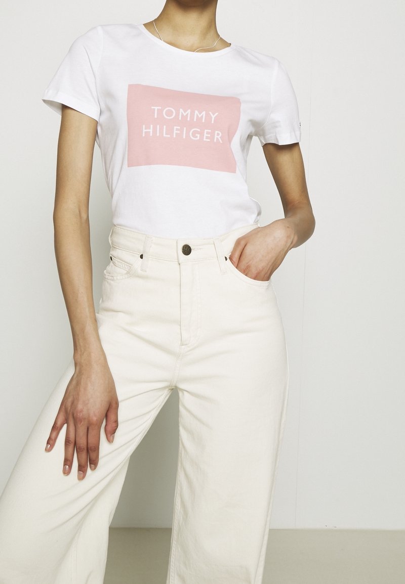 Camiseta blanca de algodón con un logo rectangular rosa que dice "TOMMY HILFIGER". Combinada con pantalones de pierna ancha color crema de cintura alta.