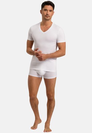 Mężczyzna stojący boso, ubrany w biały T-shirt z dekoltem w serek oraz białe bokserki, na jednolitym tle.