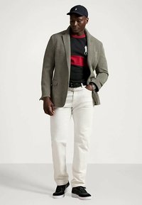 Vyras, vilkintis pilką švarką, juodai raudonais dryžuotais marškiniais, baltais kelnėmis, juodu diržu, juodu kepurėle ir juodais batais, stovi su viena ranka kišenėje.