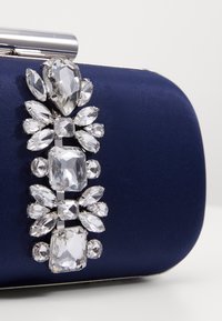 Tummansininen satiinilaukku, jossa on koristeellinen hopeanvärinen strassikoristelu, joka koostuu eri muotoisista ja kokoisista kristalleista.