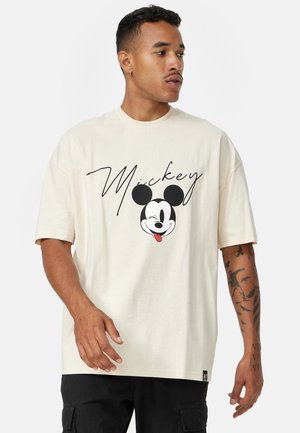 Camiseta de algodón crema con mangas cortas, que presenta un gráfico impreso de Mickey Mouse y el texto "Mickey" en cursiva negra. Ajuste clásico.
