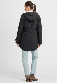 Zwarte waterdichte parka met een capuchon, een getailleerde taille en een zachte voering. Kenmerken zijn lange mouwen en een afgeronde zoom, gedragen met lichte denim en gebloemde laarzen.