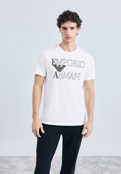 Balta kokvilnas T-krekls ar melnu "Emporio Armani" logotipu priekšpusē. Īsas piedurknes un apaļa izgriezuma kakls. Modelis valkā melnas bikses.