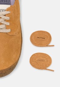 Scarpe in pelle marrone chiaro con tomaia texturizzata e lacci beige chiaro. Due lacci tondi aggiuntivi marrone chiaro accanto alla scarpa, con una finitura liscia.