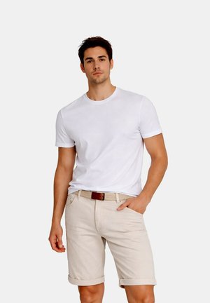Joven con camiseta blanca lisa y pantalones cortos beige remangados con un cinturón trenzado, de pie con una mano en el bolsillo frente a un fondo blanco.