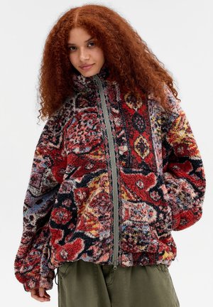 Frau mit lockigem roten Haar, die eine mehrfarbige gemusterte Fleecejacke mit Reißverschluss und grüne Hose vor einem einfarbigen hellen Hintergrund trägt.