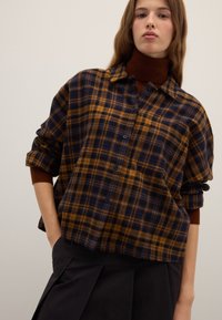 Button-up plaid shirt in navy and mustard, featuring a relaxed fit, rolled sleeves, and a classic collar. Soft fabric with a textured finish.