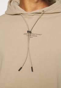 Sudadera con capucha beige con cordones de punta negra, texto de marca en negro en el pecho y una tela suave y texturizada.
