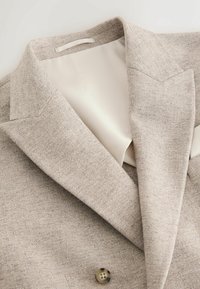 Svetlobéžové textúrované sako s výstrihom s výrezom, jedným gombíkom a hladkou krémovou podšívkou vo vnútri oblasti goliera.