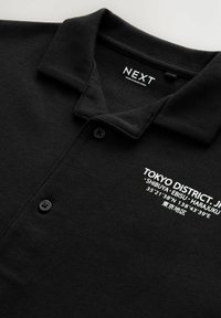 Camisa polo negra de algodón, con collar y tres botones; texto en blanco que muestra "TOKYO DISTRICT" y coordenadas.