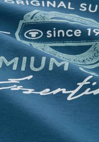 Blauwe stof met een vintage-stijl grafische print, witte tekst en een circulair logo-ontwerp. De tekst bevat "sinds 19" gevolgd door "Premium Essentials."