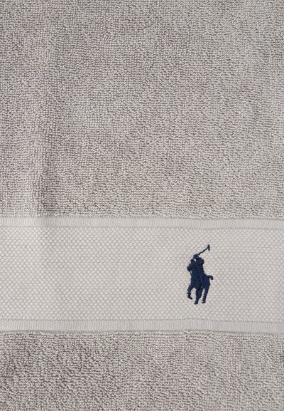 Πετσέτα σε μπεζ απόχρωση με υφή, με οριζόντια ταινία που απεικονίζει ένα σκούρο μπλε κεντημένο παίκτη πόλο πάνω σε άλογο.