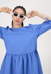 Kék textúrájú ruha, összegyűrt derékkal és puffos ujjal. A modellen fehér téglalap alakú napszemüveg és karika fülbevaló van.