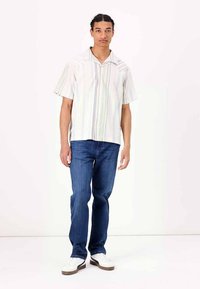 Camisa de manga corta con cuello y rayas verticales multicolores, combinada con jeans de mezclilla azules y zapatillas blancas, de pie contra un fondo liso.