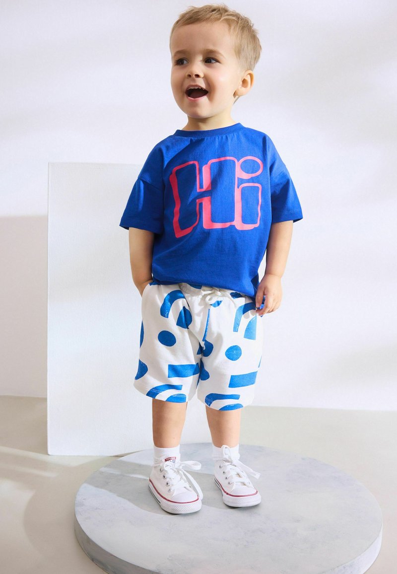 Toddler with short blonde hair wearing a blue "Hi" shirt, white shorts with blue patterns, white socks, and white sneakers standing on a round platform.