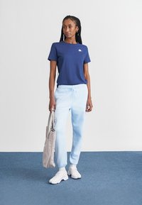 Ναυτικό μπλε T-shirt με μικρό λευκό λογότυπο, ανοιχτό μπλε gradient παντελόνι φόρμας και λευκά αθλητικά παπούτσια, κρατώντας μια ανοιχτό γκρι τσάντα ώμου.