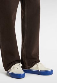 Witte canvas sneakers met blauwe rubberen zolen, gedragen met bruin, ontspannen-pasvorm broek met contrasterende stiksel langs de naden.