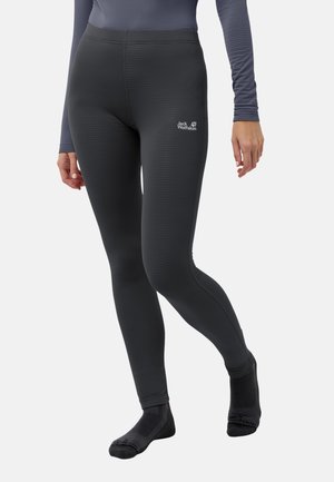 Frau trägt schwarze Jack Wolfskin Leggings und passende schwarze Socken, läuft mit einem Fuß angehoben vor einem schlichten hellen Hintergrund.