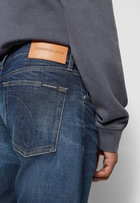 Mörkblå denimjeans med en strukturerad yta, utrustade med en beige läderlapp märkt "CALVIN KLEIN JEANS" och karakteristisk fickstickning.