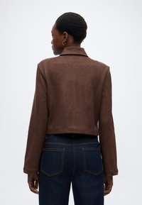 Braune Wildlederjacke in kurzer Form mit Kragen, langen Ärmeln und glatter Textur, kombiniert mit dunkelblauen Jeans mit Gesäßtaschen.