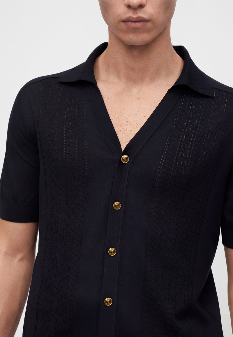 Homme portant une chemise noire à manches courtes avec des boutons décoratifs dorés et un subtil motif vertical sur le tissu.
