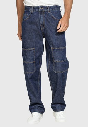 Uomo che indossa jeans cargo in denim blu con grandi tasche sulle ginocchia, una camicia bianca e scarpe da ginnastica bianche, in piedi davanti a uno sfondo monocromatico.