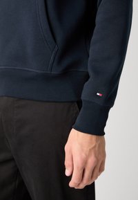 Gros plan sur le bras d'une personne portant un sweat-shirt bleu marine avec un petit logo rouge, blanc et bleu au niveau du poignet, associé à un pantalon noir.