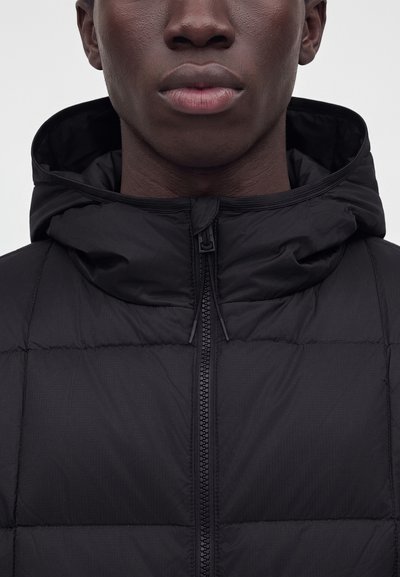 Gros plan d'une personne portant une doudoune noire zippée avec une capuche sur un fond uni.