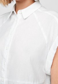 Vit linneskjorta med knappdesign, korta uppvikta ärmar och klassisk krage. Tyget verkar mjukt och texturerat.