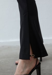 Zwarte op maat gemaakte broek met een zijsplit aan de zoom en een gouden detail op een dunne band, in combinatie met een zwarte haksandalen.